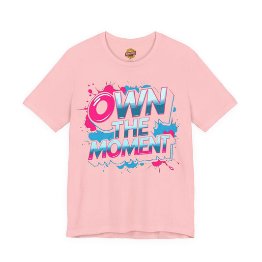 Pink t-shirt with 'OWN THE MOMENT' text and colorful design on a white background