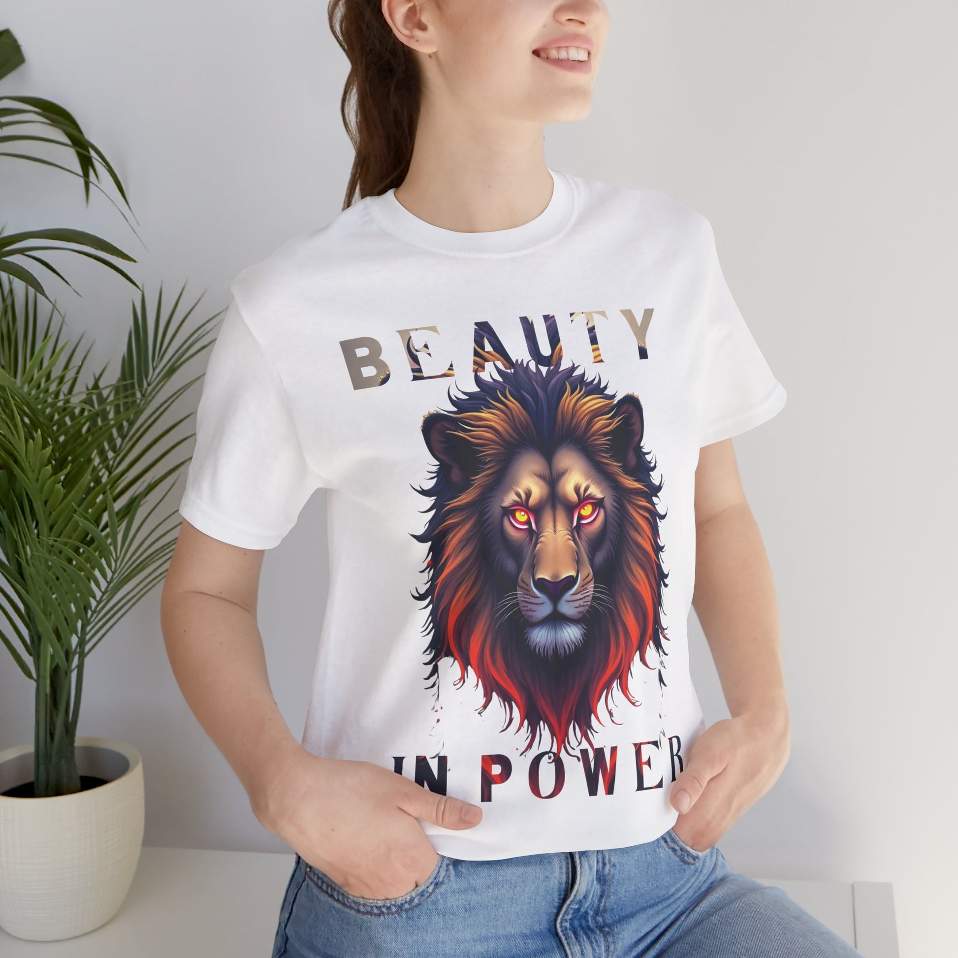 Person wearing a white t-shirt with a lion graphic and text, standing indoors next to a plant.