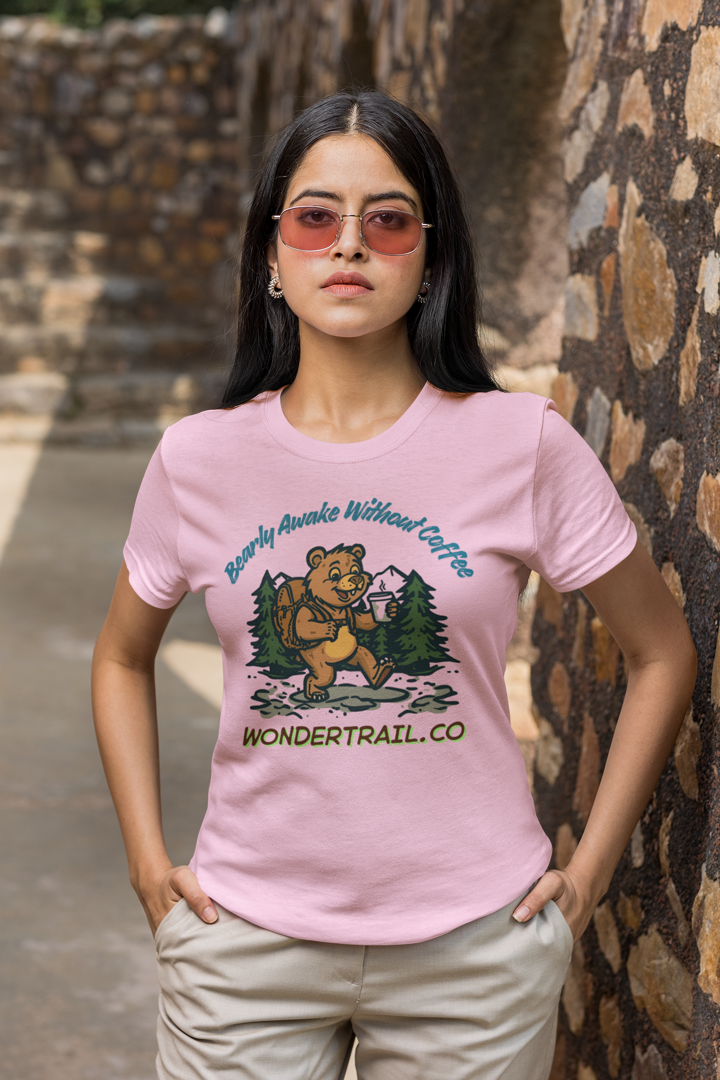 Woman wearing a pink t-shirt with a bear graphic and text, standing against a stone wall.