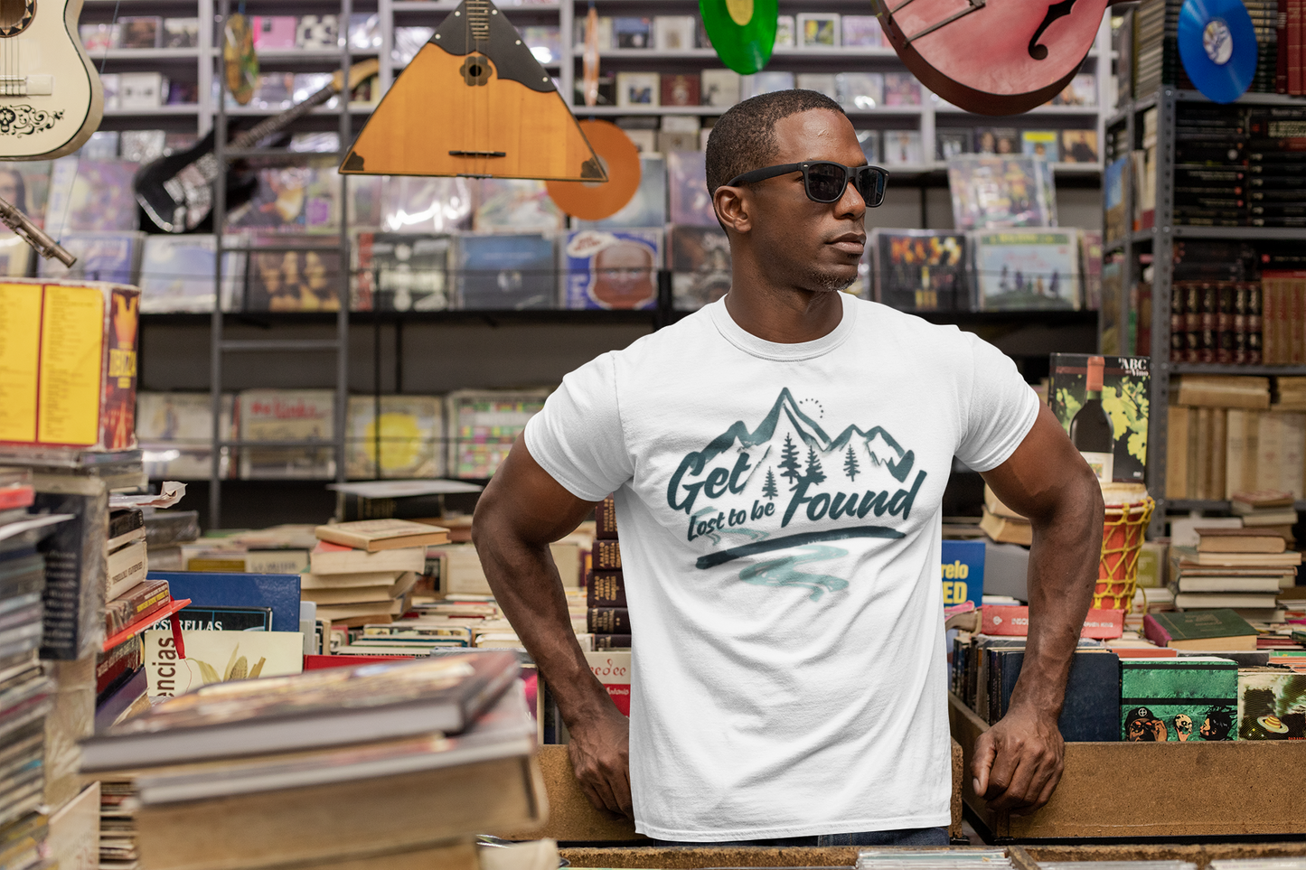 Adventure graphic T-shirt with mountain and forest design, slogan ‘Get lost to be found,’ styled in a bookstore with records and guitars — casual outdoor fashion.
